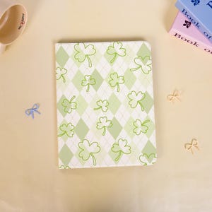 May include: A rectangular notebook featuring a light green and white argyle diamond pattern overlaid with green shamrock illustrations. The cover has a smooth finish and rounded corners, presenting a consistent repeating clover motif throughout.