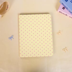 May include: A rectangular notebook with a pale yellow cover featuring a repeating pattern of small pink polka dots. The notebook is surrounded by a light blue mug, two stacked books with visible text, and three small bow-shaped decorative clips.