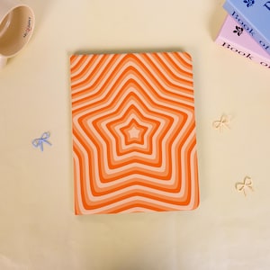 Könnte beinhalten: Ein Notizbuch mit einem Sternmuster in Orange- und Weißtönen. Das Cover zeigt konzentrische Sternformen, die einen Retro- und psychedelischen Effekt erzeugen. Das Notizbuch ist rechteckig und liegt flach auf einer hellen Oberfläche.