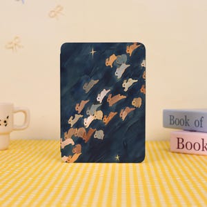 Może przedstawiać: Prostokątne etui na tablet z fantazyjną ilustracją licznych królików w różnych odcieniach brązu i bieli na ciemnoniebieskim tle. Projekt zawiera małe gwiazdki, tworząc efekt nocnego nieba. Etui znajduje się na żółtej, w kratkę powierzchni.