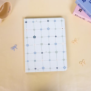 May include: A light blue notebook with a floral grid pattern. The notebook features a grid of thin gray lines with small blue and gray flowers at the intersections. The cover is surrounded by small decorative bows and books.