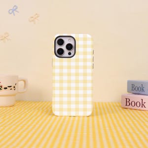 May include: A yellow and white checked phone case. The case has a square pattern and a black camera lens area. The phone case is on a yellow checked surface.