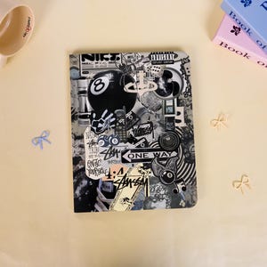 Könnte beinhalten: Ein rechteckiges Notizbuch mit einer Collage aus schwarz-weißen Aufklebern, darunter eine Billardkugel mit der Nummer acht, eine Spinne, Würfel, ein Mobiltelefon, ein Einbahnstraßenschild sowie Text mit Parental Advisory Explicit Content, Stussy.