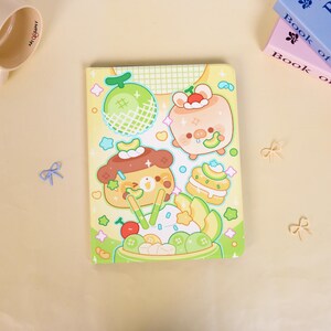 Op de afbeelding: Een rechthoekig notitieboek met een lichtgele kaft met een kleurrijke illustratie van een meloen, een varkentje, een beertje en diverse desserts. Het ontwerp bevat groene, bruine en roze accenten met kleine ster- en sprankelmotieven verspreid over het oppervlak.