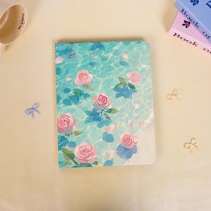 May include: A rectangular notebook featuring a light blue and turquoise water surface pattern with floating pink roses and green leaves. The cover design includes scattered rose petals across the rippling water effect, creating a serene visual.