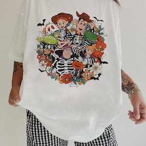 May include: A white t-shirt featuring a colorful graphic print of various animated characters dressed in skeleton costumes. The design includes orange flowers, spider webs, bats, and pumpkins, creating a festive Halloween-themed illustration.