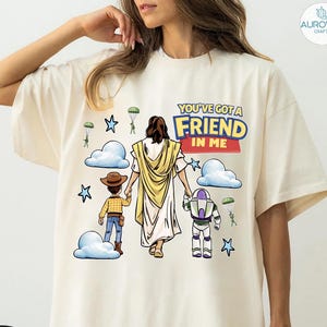 May include: Ivory-colored t-shirt featuring a graphic with the text "YOU'VE GOT A FRIEND IN ME." The design includes illustrations of Jesus, Woody, and Buzz Lightyear, with clouds, stars, and parachutes. The shirt has a relaxed fit.