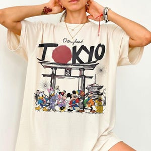 Puede incluir: Camiseta color crema con un dise&ntilde;o de Disneyland Tokyo. El dise&ntilde;o incluye la palabra "Tokyo" en negrita, un c&iacute;rculo rojo y una representaci&oacute;n de personajes de Disney frente a una puerta y una pagoda japonesas.