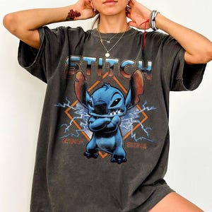 Puede incluir: Camiseta gris oscuro con un personaje de dibujos animados azul y la palabra "STITCH" en una fuente retro. El dise&ntilde;o incluye rayos y un marco en forma de diamante, creando una est&eacute;tica vintage. La camiseta es de gran tama&ntilde;o y tiene mangas cortas.