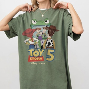 K&ouml;nnte beinhalten: Olivgr&uuml;nes T-Shirt mit einem Grafikdesign aus dem Film Toy Story 5. Das Design zeigt Charaktere wie Woody und Jessie sowie einen Frosch, mit dem Text "TOY STORY 5" in einer bunten Schriftart. Das Disney Pixar-Logo befindet sich unten.
