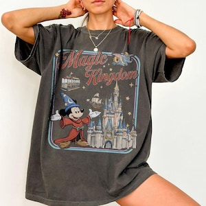 May include: Dark gray t-shirt featuring a vintage-style graphic with the words "Magic Kingdom." The design includes a castle, Mickey Mouse in a wizard outfit, and other theme park elements. The shirt has short sleeves and a relaxed fit.
