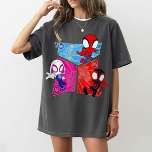 May include: Dark gray t-shirt featuring a colorful graphic of Spider-Man characters. The design includes Spider-Man, Spider-Gwen, and Miles Morales in a cartoon style. The shirt has a crew neck and short sleeves.