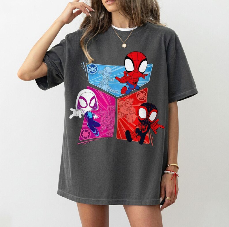 May include: Dark gray t-shirt featuring a colorful graphic of Spider-Man characters. The design includes Spider-Man, Spider-Gwen, and Miles Morales in a cartoon style. The shirt has a crew neck and short sleeves.