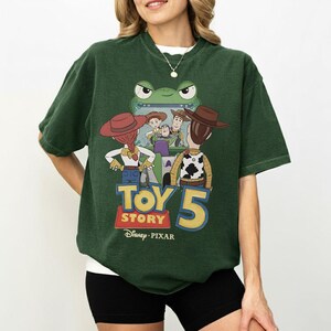 K&ouml;nnte beinhalten: Dunkelgr&uuml;nes T-Shirt mit einer Grafik aus dem Film Toy Story 5. Das Design zeigt Woody, Buzz Lightyear, Jessie und andere Charaktere. Das T-Shirt zeigt den Text "Toy Story 5" und das Disney Pixar-Logo.