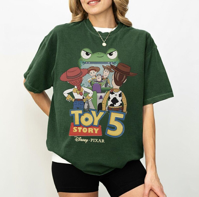 K&ouml;nnte beinhalten: Dunkelgr&uuml;nes T-Shirt mit einer Grafik aus dem Film Toy Story 5. Das Design zeigt Woody, Buzz Lightyear, Jessie und andere Charaktere. Das T-Shirt zeigt den Text "Toy Story 5" und das Disney Pixar-Logo.