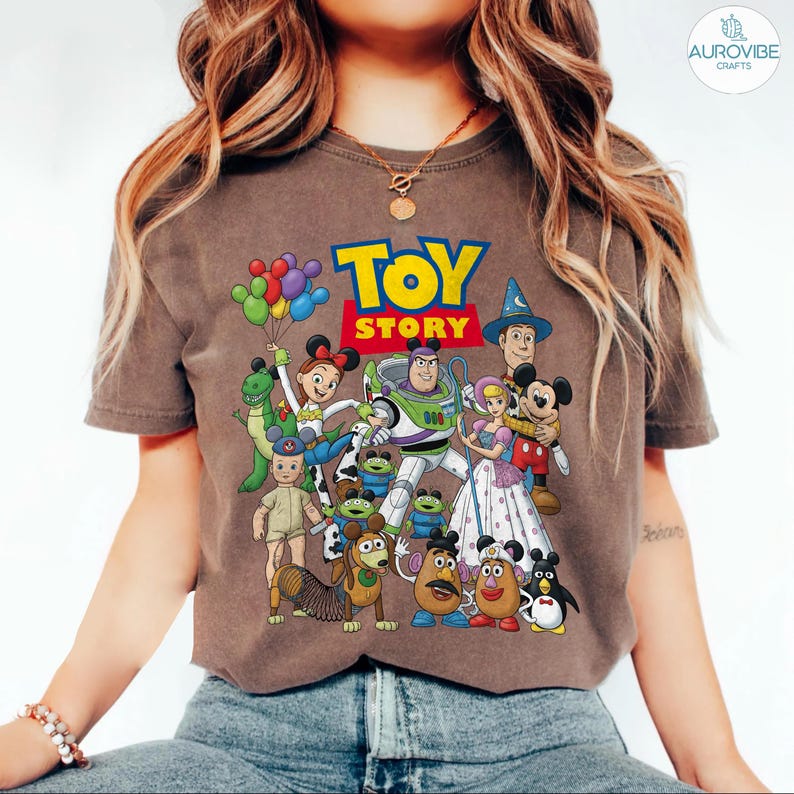 Op de afbeelding: Een bruin t-shirt met een kleurrijke afbeelding van personages uit de film "Toy Story". Het ontwerp bevat Buzz Lightyear, Woody, Mickey en Minnie Mouse en andere personages. De woorden "Toy Story" zijn gedrukt in een vet, kleurrijk lettertype.