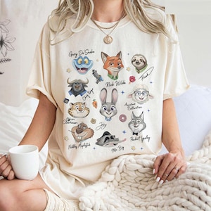 Op de afbeelding: Cr&egrave;mekleurig T-shirt met cartoonillustraties van personages uit de film Zootopia. Het shirt bevat afbeeldingen van Judy Hopps, Nick Wilde en andere personages, met hun namen eronder geschreven. Het shirt is een casual kledingstuk.