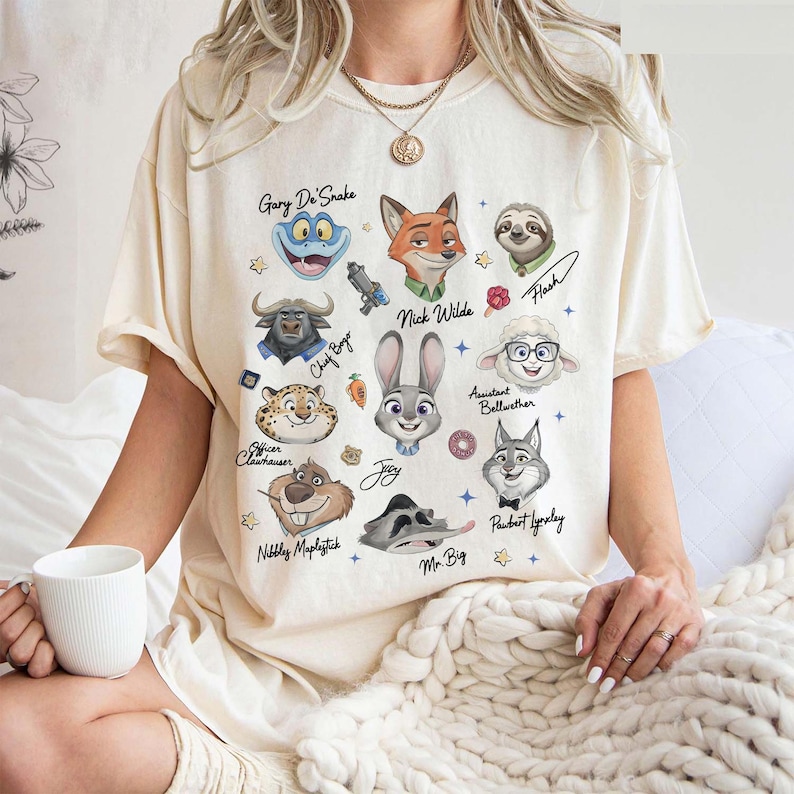 Op de afbeelding: Cr&egrave;mekleurig T-shirt met cartoonillustraties van personages uit de film Zootopia. Het shirt bevat afbeeldingen van Judy Hopps, Nick Wilde en andere personages, met hun namen eronder geschreven. Het shirt is een casual kledingstuk.