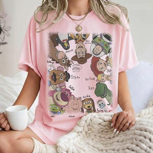 K&ouml;nnte beinhalten: Hellrosa T-Shirt mit einem farbenfrohen Aufdruck verschiedener Toy Story-Charaktere, darunter Woody, Buzz Lightyear und Rex. Die Namen der Charaktere sind in einer verspielten Schrift geschrieben. Ein l&auml;ssiger, bequemer Stil.