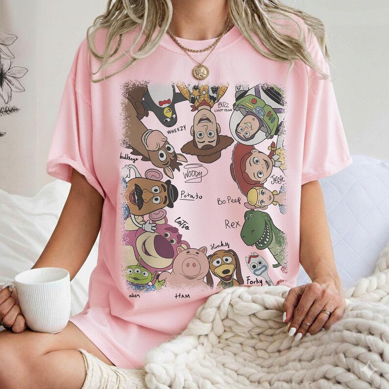 K&ouml;nnte beinhalten: Hellrosa T-Shirt mit einem farbenfrohen Aufdruck verschiedener Toy Story-Charaktere, darunter Woody, Buzz Lightyear und Rex. Die Namen der Charaktere sind in einer verspielten Schrift geschrieben. Ein l&auml;ssiger, bequemer Stil.