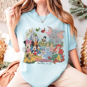 Puede incluir: Camiseta azul claro con un gr&aacute;fico colorido de personajes de Disney, incluyendo Mickey Mouse, Minnie Mouse y Dumbo. El texto "Magic Kingdom Walt Disney World" est&aacute; impreso debajo de los personajes.