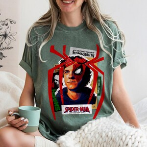 Op de afbeelding: Groen t-shirt met een Spider-Man-afbeelding. Het ontwerp bevat een portret van Spider-Man, een rood spinnenweb en de tekst "SPIDER-MAN BRAND NEW DAY" en "WITH GREAT POWER COMES GREAT RESPONSIBILITY".