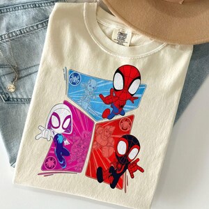 May include: Cream-colored t-shirt featuring a graphic of Spider-Man characters in red, blue, and pink diamond shapes. The shirt has a Comfort Colors tag and is laid on top of blue jeans and a tan hat.