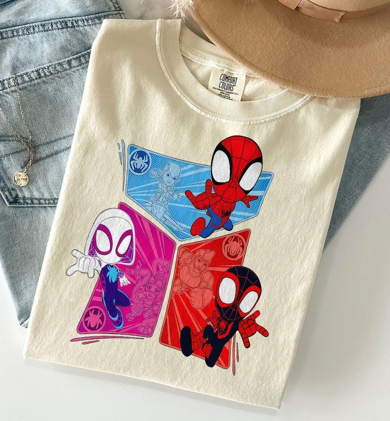 May include: Cream-colored t-shirt featuring a graphic of Spider-Man characters in red, blue, and pink diamond shapes. The shirt has a Comfort Colors tag and is laid on top of blue jeans and a tan hat.