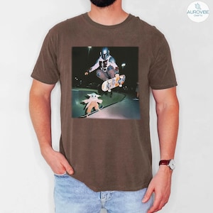Puede incluir: Camiseta marr&oacute;n con un gr&aacute;fico de un skater y un personaje de una popular serie de ciencia ficci&oacute;n. El gr&aacute;fico muestra al skater en el aire, realizando un truco, con otro personaje patinando debajo.