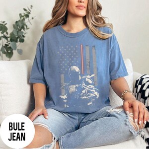 May include: Blue t-shirt featuring a distressed American flag design with a character graphic. The shirt has short sleeves and a crew neck. The text "BULE JEAN" is in a white circle at the bottom left.
