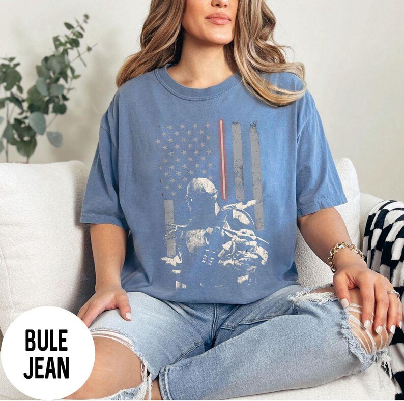 May include: Blue t-shirt featuring a distressed American flag design with a character graphic. The shirt has short sleeves and a crew neck. The text "BULE JEAN" is in a white circle at the bottom left.