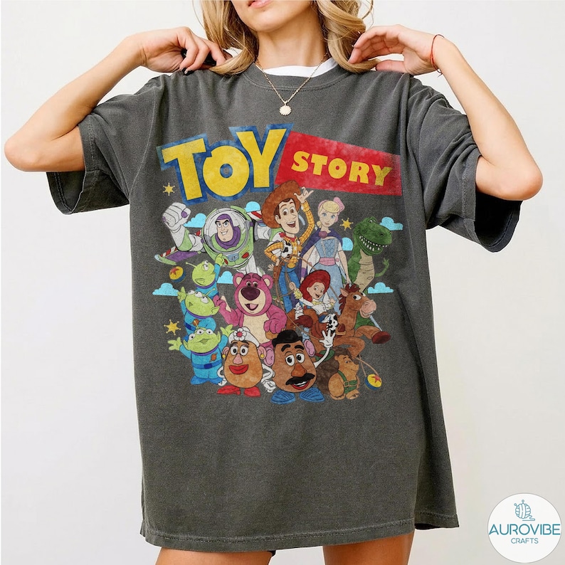 May include: Dark gray t-shirt featuring the words "TOY STORY" in yellow and red, with a graphic of characters from the animated film. The design includes Woody, Buzz Lightyear, and other characters. The shirt has short sleeves.