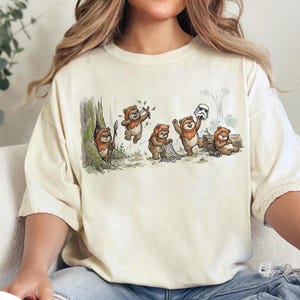 May include: Cream-colored t-shirt featuring a whimsical illustration of Ewoks in a forest setting. The design shows several Ewoks engaged in various activities, including hunting and holding a Stormtrooper helmet. The artwork is detailed and colorful.