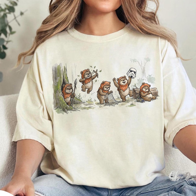 May include: Cream-colored t-shirt featuring a whimsical illustration of Ewoks in a forest setting. The design shows several Ewoks engaged in various activities, including hunting and holding a Stormtrooper helmet. The artwork is detailed and colorful.
