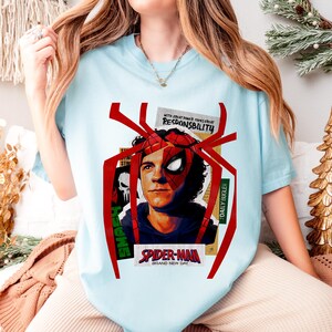 Op de afbeelding: Lichtblauw T-shirt met een Spider-Man graphic en de tekst "Spider-Man Brand New Day". Het ontwerp bevat de woorden "With great power comes great responsibility" en "Daily Bugle". Het shirt heeft een ronde hals.