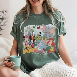 Puede incluir: Camiseta verde con un gr&aacute;fico colorido de personajes de Disney, incluyendo Mickey Mouse, Minnie Mouse, Donald Duck y Ariel. El gr&aacute;fico incluye el texto "Magic Kingdom, Walt Disney World".