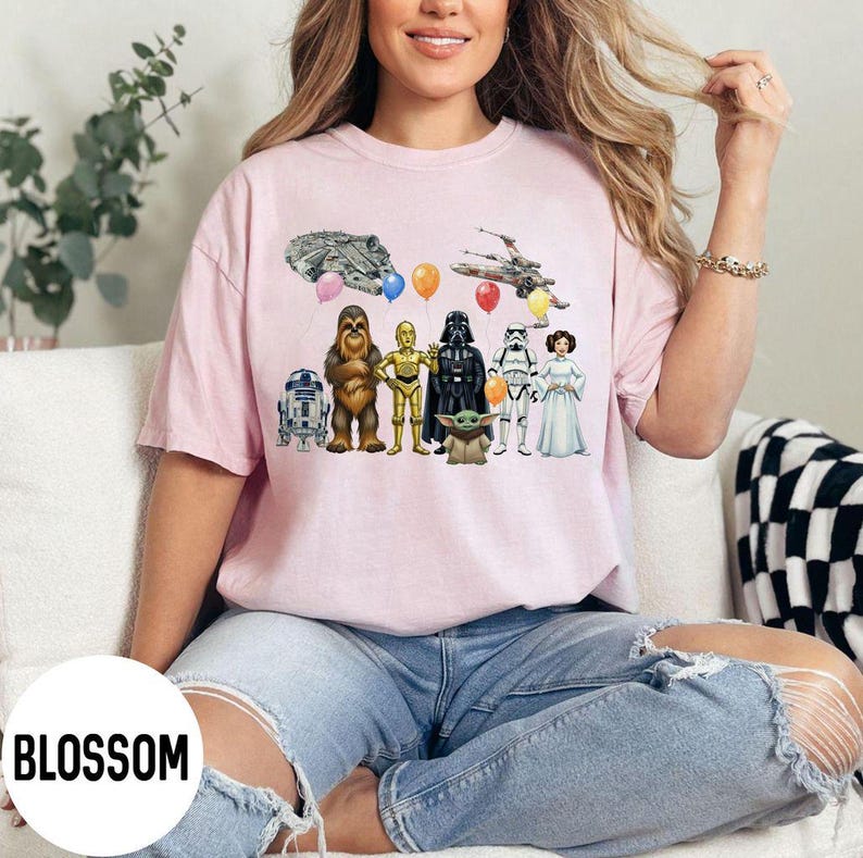 Peut inclure: T-shirt rose clair avec un motif Star Wars. Le design comprend des illustrations de personnages, de vaisseaux spatiaux et de ballons. Le mot "BLOSSOM" est imprim&eacute; dans un cercle blanc en bas &agrave; gauche.
