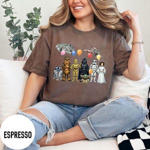 Peut inclure: T-shirt couleur expresso avec un motif Star Wars. Le design comprend divers personnages, des vaisseaux spatiaux et des ballons. Le mot "ESPRESSO" est imprim&eacute; en blanc en bas &agrave; gauche du t-shirt.