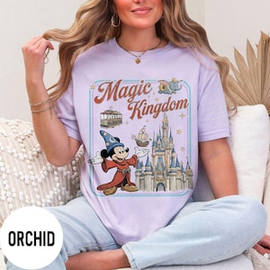 May include: A light purple t-shirt featuring a graphic of Mickey Mouse in a sorcerer's outfit, a castle, and the words "Magic Kingdom." The design includes a trolley, a ship, and Dumbo the elephant. The word "ORCHID" is printed at the bottom.