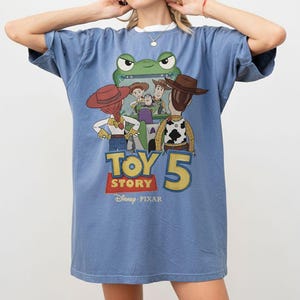 K&ouml;nnte beinhalten: Hellblaues T-Shirt mit einem Toy Story 5-Motiv. Das Design zeigt Charaktere wie Woody und Buzz Lightyear sowie den Filmtitel und das Disney Pixar-Logo. Das T-Shirt hat kurze &Auml;rmel und eine lockere Passform.
