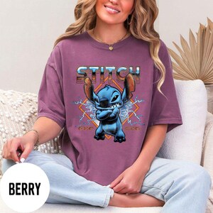 Puede incluir: Camiseta color bayas con un gr&aacute;fico de Stitch de Lilo & Stitch. El personaje azul est&aacute; frente a un dise&ntilde;o en forma de diamante con rayos. La palabra "STITCH" est&aacute; impresa encima del personaje en una fuente met&aacute;lica.