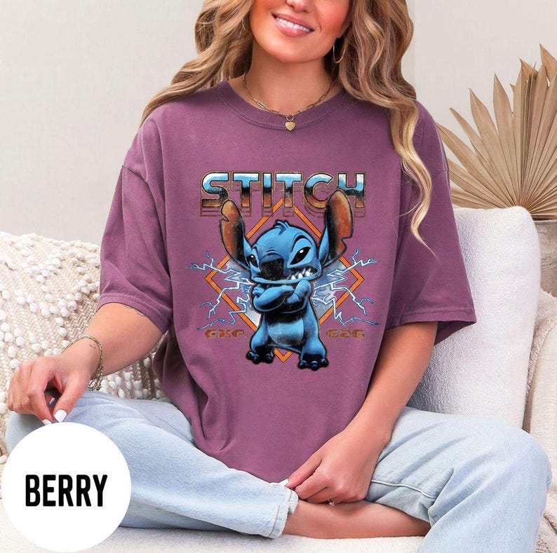 Puede incluir: Camiseta color bayas con un gr&aacute;fico de Stitch de Lilo & Stitch. El personaje azul est&aacute; frente a un dise&ntilde;o en forma de diamante con rayos. La palabra "STITCH" est&aacute; impresa encima del personaje en una fuente met&aacute;lica.