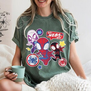 Puede incluir: Camiseta verde con un dise&ntilde;o de dibujos animados de tres personajes de Spider-Man. El dise&ntilde;o incluye a Spider-Man, Spider-Gwen y un Spider-Man negro, junto con el texto "GO WEBS - GO!". La camiseta tiene cuello redondo y mangas cortas.
