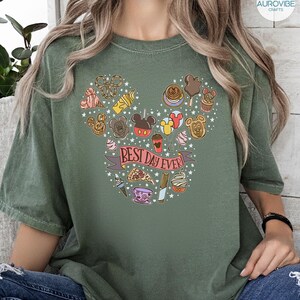May include: An olive green t-shirt featuring a graphic print of various snacks and treats arranged in the shape of a mouse head silhouette. A pink banner across the center contains the text "BEST DAY EVER!" surrounded by small white stars.