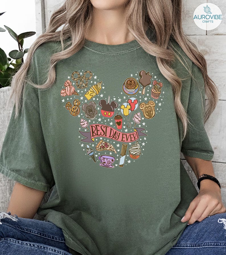 May include: An olive green t-shirt featuring a graphic print of various snacks and treats arranged in the shape of a mouse head silhouette. A pink banner across the center contains the text "BEST DAY EVER!" surrounded by small white stars.