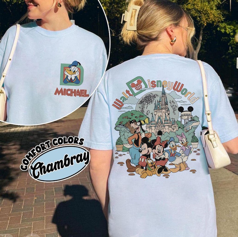 May include: Light blue Comfort Colors Chambray t-shirt featuring a Donald Duck graphic with the name "Michael" on the front. The back of the shirt displays a colorful Disney World design with Mickey Mouse and friends.