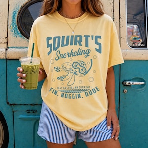 May include: Yellow t-shirt with the text "Squirt's Snorkeling" and a turtle graphic. The shirt also features the phrases "Fin, Noggin, Dude" and "East Australian Current" in blue. The person is wearing blue and white striped shorts.