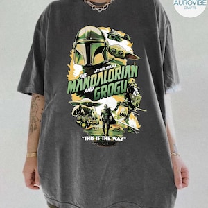 Pode incluir: T-shirt cinzenta escura com um design gr&aacute;fico da s&eacute;rie Star Wars, "The Mandalorian and Grogu". O design inclui imagens do capacete Mandaloriano, Grogu e outros personagens, com a frase "THIS IS THE WAY."