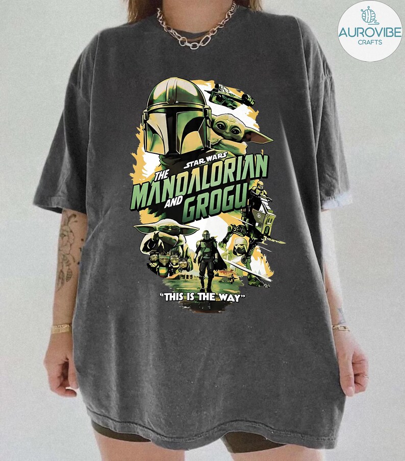 Pode incluir: T-shirt cinzenta escura com um design gr&aacute;fico da s&eacute;rie Star Wars, "The Mandalorian and Grogu". O design inclui imagens do capacete Mandaloriano, Grogu e outros personagens, com a frase "THIS IS THE WAY."
