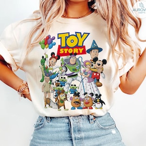 Op de afbeelding: Cr&egrave;mekleurig T-shirt met een kleurrijke afbeelding van personages uit de animatiefilm "Toy Story". Het ontwerp bevat de titel van de film in vette letters en verschillende personages zoals Buzz Lightyear en Woody.